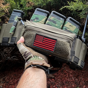 May include: An olive green tactical chest rig with multiple magazine pouches and an American flag patch. The rig holds several black magazines with camouflage wraps. A small camouflage-patterned item is attached to the rig.