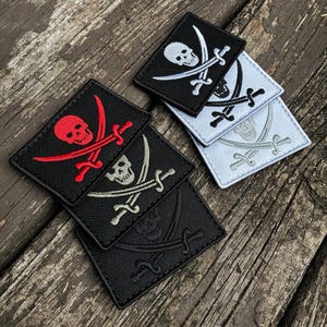 May include: A collection of embroidered pirate patches in various colors. The patches feature a skull and crossbones design with crossed swords. Colors include black, red, white, and gray. The patches are square-shaped and have a textured appearance.