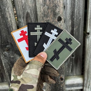 May include: Four rectangular embroidered patches featuring a double-barred cross symbol. The patches are arranged in a fan shape, showcasing a white patch with a red cross, a black patch with an olive cross, a black patch with a white cross, and an olive patch with a black cross.