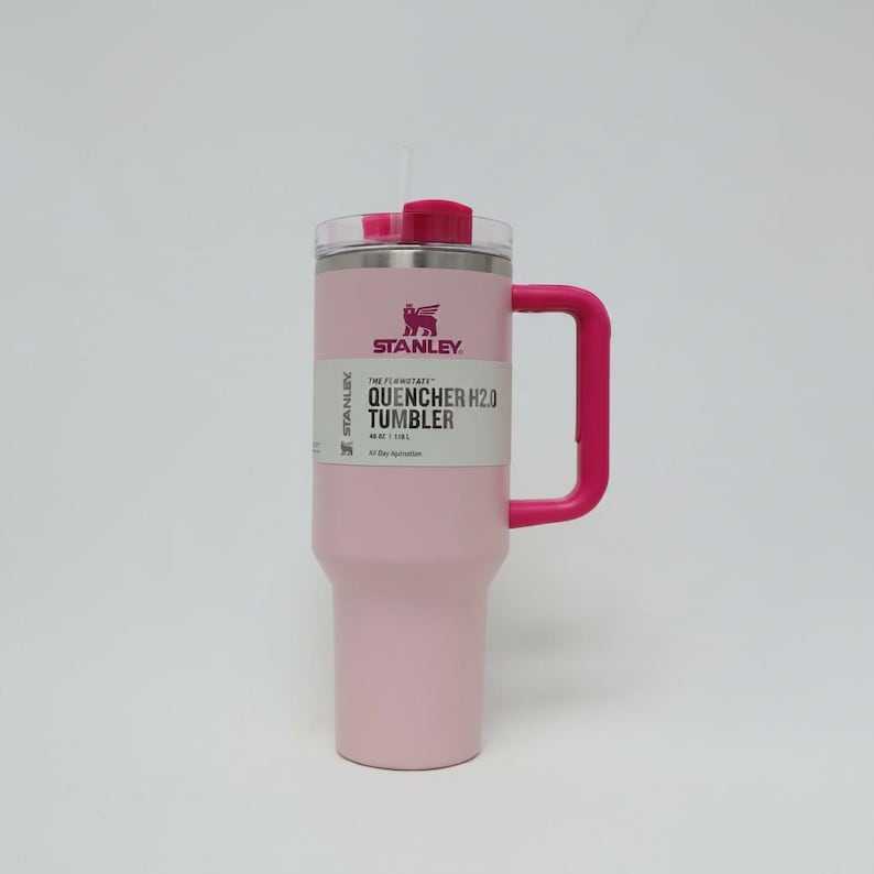 May include: A pink Stanley Quencher H2.0 Tumbler with a pink handle and lid. The tumbler has a white label with black text and a logo. It is designed to hold 40 oz (1.18 L) of liquid.