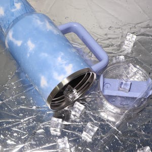 K&ouml;nnte beinhalten: Ein hellblauer isolierter Becher mit wei&szlig;em Wolkenmuster, passendem Griff und transparentem Deckel. Der Edelstahlrand ist sichtbar. Eisw&uuml;rfel und Wassertropfen umgeben den Becher, was auf ein kaltes Getr&auml;nk hindeutet.