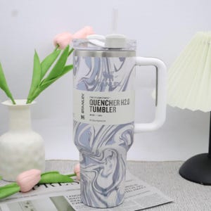 May include: A stainless steel Stanley Quencher H2.0 tumbler with a white handle and straw. The tumbler features a swirling blue and white pattern and a white lid. The label reads "QUENCHER H2.0 TUMBLER" and "40 oz | 1.18L".