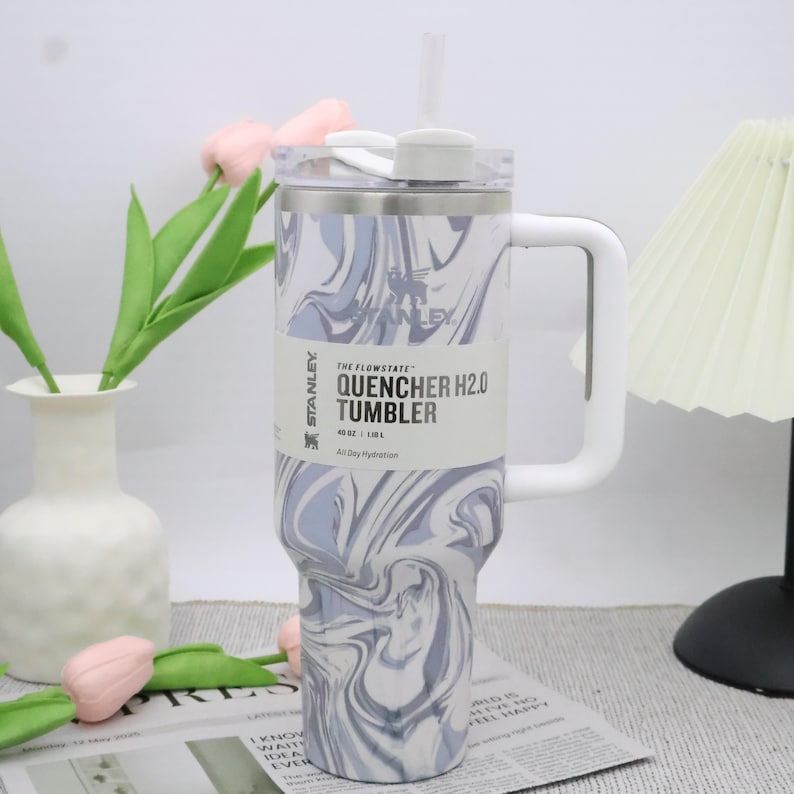 May include: A stainless steel Stanley Quencher H2.0 tumbler with a white handle and straw. The tumbler features a swirling blue and white pattern and a white lid. The label reads "QUENCHER H2.0 TUMBLER" and "40 oz | 1.18L".