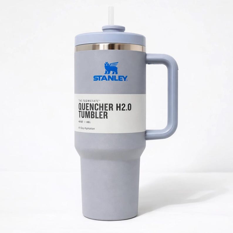 May include: A gray Stanley Quencher H2.0 Tumbler with a light blue lid, handle, and straw. The tumbler has a silver band and the Stanley logo in blue. The text "QUENCHER H2.0 TUMBLER" is printed on the front. It holds 40 oz (1.18L).
