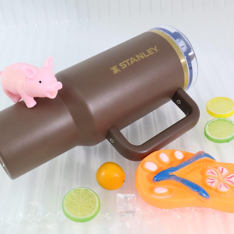 May include: A brown Stanley tumbler with a handle, a clear lid, and gold accents. A pink pig figurine, orange flip-flop, and lemon and lime slices are next to the tumbler. The tumbler has the word "STANLEY" printed on it.