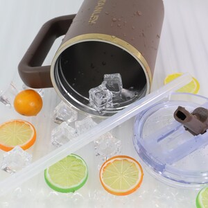 May include: A brown insulated tumbler with a handle, gold trim, and a clear lid with a straw. The tumbler is partially filled with ice cubes. Decorative orange and lemon slices are scattered around the tumbler.