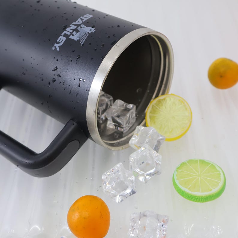 May include: A black Stanley insulated mug with a handle, silver rim, and the Stanley logo. Ice cubes and slices of lemon and lime are scattered around the mug, along with small oranges. The mug is partially filled with ice.