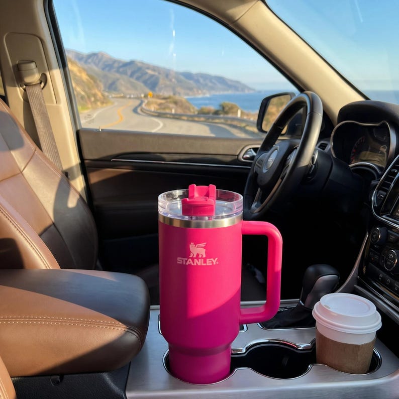 Peut inclure: Un mug de voyage Stanley rose vif avec une poign&eacute;e et un couvercle transparent est plac&eacute; dans le porte-gobelet d'une voiture. Le mug porte le logo Stanley en blanc. Une petite tasse &agrave; caf&eacute; avec un couvercle blanc est &agrave; c&ocirc;t&eacute; du mug.