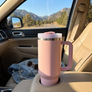 K&ouml;nnte beinhalten: Ein gro&szlig;er, rosa isolierter Reisebecher mit Griff und Deckel, in einem Getr&auml;nkehalter im Auto. Der Becher ist zylindrisch mit hellrosa Griff und Deckel. Im Hintergrund Berge und eine Stra&szlig;e.