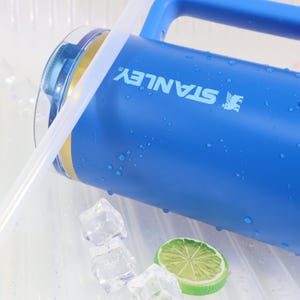 May include: A blue Stanley water bottle with a handle and a clear straw. The bottle has the Stanley logo printed on it in white. The bottle is wet with water droplets. Ice cubes and a lime slice are in the background.