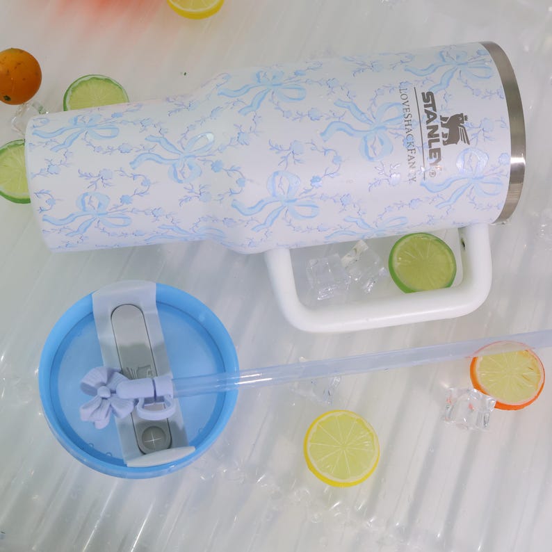 K&ouml;nnte beinhalten: Ein wei&szlig;er Stanley-Becher mit blauem Blumenmuster und Griff. Der Becher hat einen blauen Deckel und einen durchsichtigen Strohhalm. Der Text "STANLEY" und "LOVESHAKEFAN" ist sichtbar. Der Becher ist von Eis, Zitronen- und Orangenscheiben umgeben.
