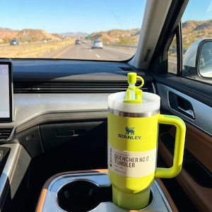 Op de afbeelding: Een felgele Stanley Quencher H2.0 Tumbler met een handvat en een wit deksel staat in de bekerhouder van een auto. De tumbler heeft het Stanley-logo en productinformatie. De achtergrond toont een snelweg en bergen.