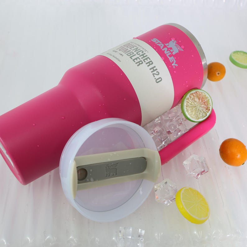 Pu&ograve; includere: Un bicchiere Stanley Quencher H2.0 rosa con una fascia bianca e il nome del marchio. Il coperchio &egrave; rimosso, rivelando l'interno. Fette decorative di agrumi e cubetti di ghiaccio sono sparsi intorno al bicchiere.