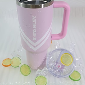 May include: A pink insulated tumbler with a handle and a clear lid. The tumbler has white diagonal stripes and the brand name "STANLEY" printed on the side. The lid has a straw and a lemon slice. Ice cubes and fruit slices surround the tumbler.
