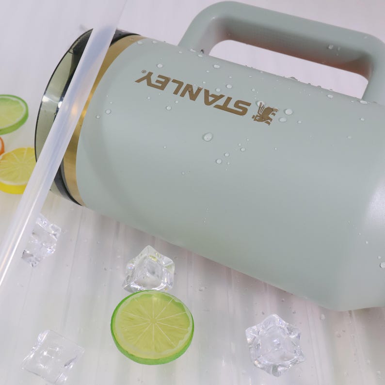 Op de afbeelding: Een lichtgroene Stanley waterkan met een handvat en een rietje. Het Stanley-logo is goudkleurig gedrukt. De kan is omgeven door ijsblokjes en limoenschijfjes. De kan is ontworpen om drankjes koud te houden.