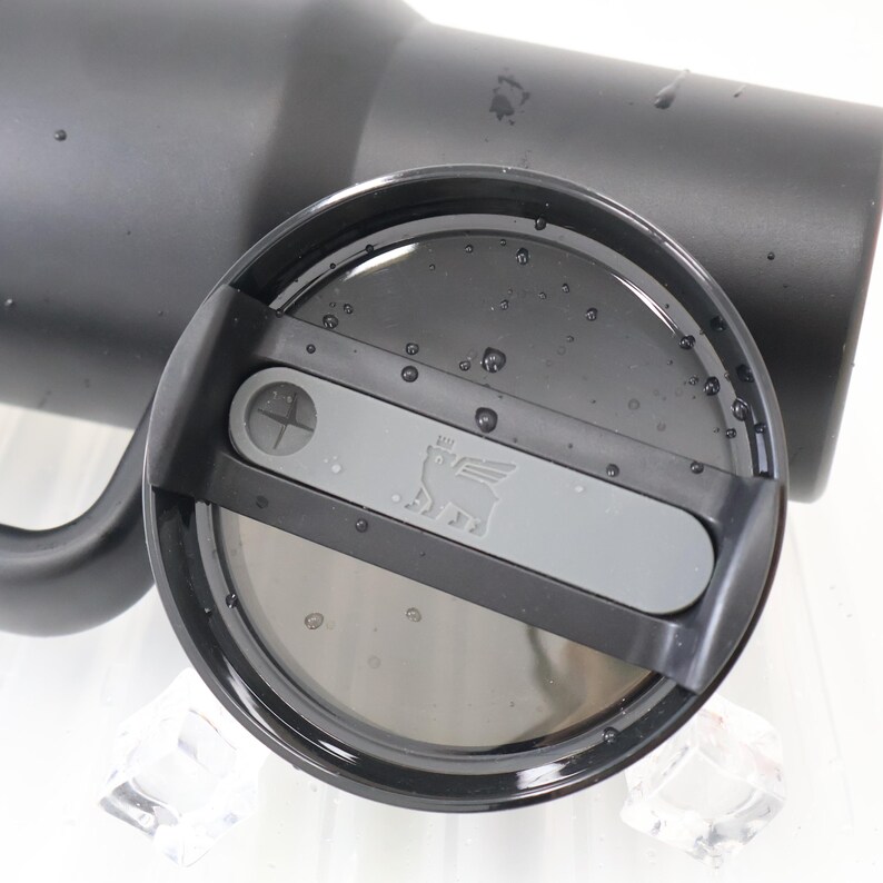 May include: A black insulated tumbler with a matching lid. The lid features a gray slider with the Stanley logo and a drinking hole. Water droplets are visible on the lid's surface. The tumbler is designed to keep beverages cold or hot.