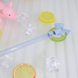May include: A light blue plastic drink dispenser spigot with a decorative handle, resting on a green lime slice. The image also includes yellow lemon slices, clear ice cubes, and a pink pig figurine, suggesting a refreshing beverage theme.