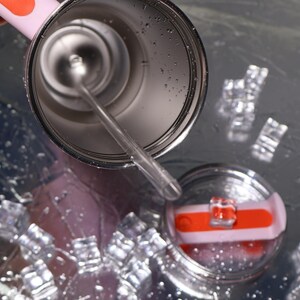May include: A pink and silver insulated tumbler with a clear straw and a matching lid. The lid has an orange accent. The tumbler is partially filled with liquid, and ice cubes are scattered around it.