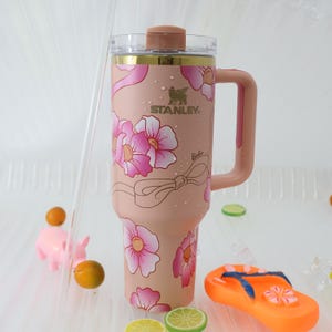 K&ouml;nnte beinhalten: Ein rosa Stanley-Becher mit floralem Design und dem Wort "Barbie". Der Becher hat einen Griff und einen transparenten Deckel. Die Blumen sind rosa und wei&szlig;. Der Becher ist mit einer Schleife verziert.