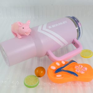 May include: A pink Stanley tumbler with a handle and white chevron design. A small pink pig figurine sits on top. Also, a slice of lemon, a small orange fruit, a lime slice, and an orange and blue flip-flop are next to the tumbler.