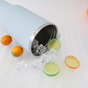 K&ouml;nnte beinhalten: Ein hellblaues, isoliertes Trinkglas mit silbernem Rand, Eisw&uuml;rfeln und Zitrusfruchtscheiben. Zwei Orangen und zwei Limettenscheiben sind um das Trinkglas herum verstreut. Das Trinkglas ist umgekippt, wobei Eisw&uuml;rfel herausfallen.