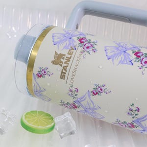 May include: A cream-colored Stanley water jug with a floral and bow pattern. The jug has a light blue handle and lid. The design includes the text "STANLEY" and "LOVESHACKFANCY". A lime slice and ice cubes are in the foreground.