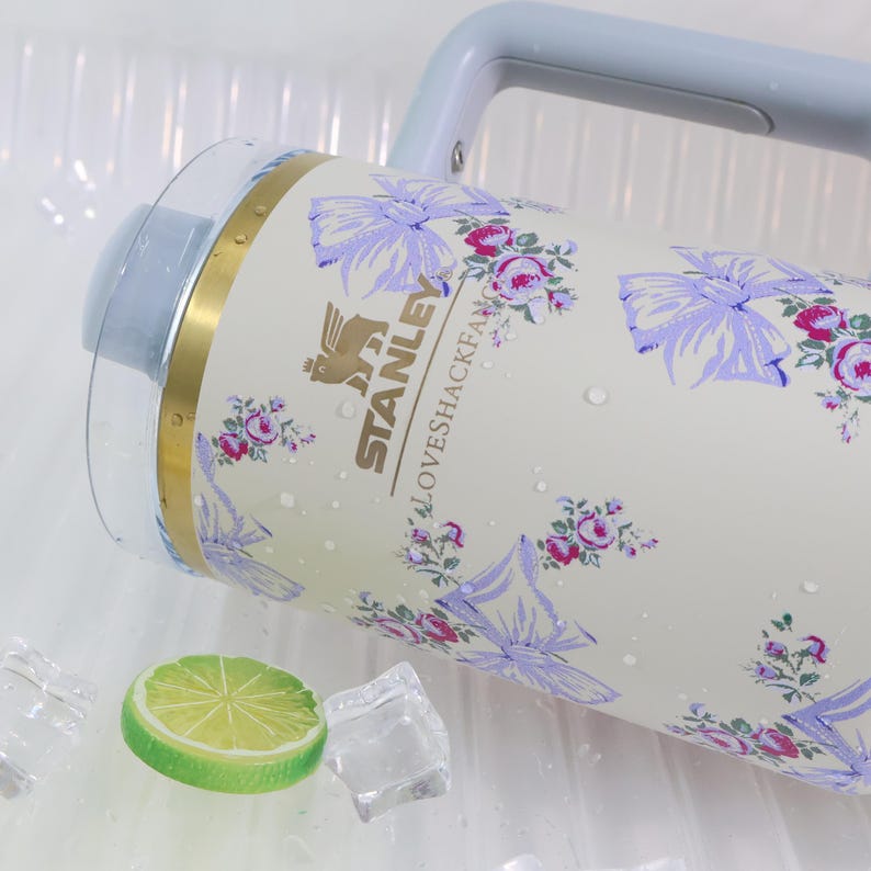 May include: A cream-colored Stanley water jug with a floral and bow pattern. The jug has a light blue handle and lid. The design includes the text "STANLEY" and "LOVESHACKFANCY". A lime slice and ice cubes are in the foreground.