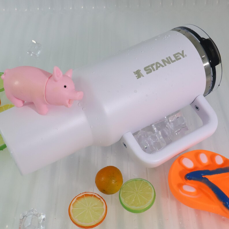 May include: A white Stanley water bottle with a handle, ice cubes, and a pink pig figurine. The bottle has the brand name printed on it. Decorative orange and lime slices and a flip-flop are also present.