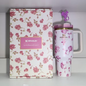 Pode incluir: Um copo Stanley branco e uma caixa a condizer, ambos adornados com um padr&atilde;o de rosas cor-de-rosa. O copo tem uma pega e um la&ccedil;o roxo decorativo na tampa. A caixa apresenta o log&oacute;tipo "Stanley LovesShackFancy".