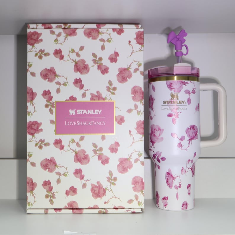 Pode incluir: Um copo Stanley branco e uma caixa a condizer, ambos adornados com um padr&atilde;o de rosas cor-de-rosa. O copo tem uma pega e um la&ccedil;o roxo decorativo na tampa. A caixa apresenta o log&oacute;tipo "Stanley LovesShackFancy".