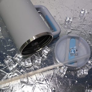 May include: A gray insulated tumbler with a handle and a lid, featuring a light blue accent. The lid has a straw and a light blue slider. The tumbler is surrounded by ice cubes and water droplets.