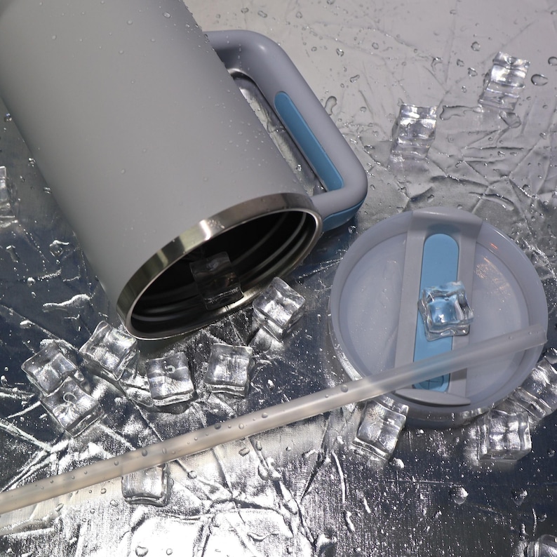 May include: A gray insulated tumbler with a handle and a lid, featuring a light blue accent. The lid has a straw and a light blue slider. The tumbler is surrounded by ice cubes and water droplets.