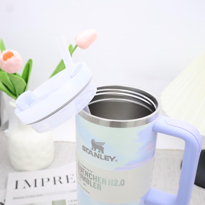 May include: A Stanley Quencher H2.0 tumbler with a light blue handle and lid. The tumbler features a pastel colour scheme with a white and light blue pattern. The lid is open, revealing the interior. The tumbler has a straw.