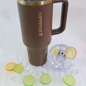 May include: A brown Stanley travel mug with a gold rim and handle. The mug is next to a clear lid with ice and citrus slices. The Stanley logo is visible on the side. Decorative ice cubes and citrus slices are scattered around.