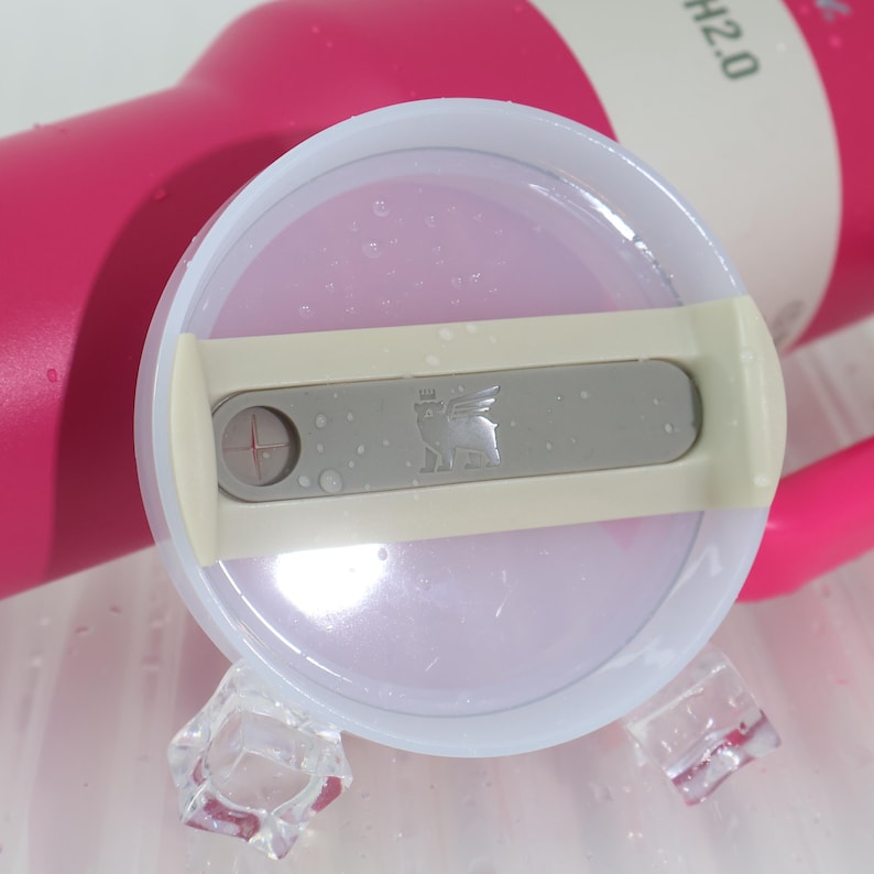 Pu&ograve; includere: Primo piano di un coperchio di bottiglia d'acqua rosa con un cursore grigio e un logo. Il coperchio &egrave; trasparente con un bordo bianco e un cursore grigio con un logo. La bottiglia &egrave; rosa e ha il testo "H2.0". Cubetti di ghiaccio in primo piano.