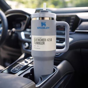 May include: A gray Stanley Quencher H2.0 tumbler with a handle and straw, resting in a car's cup holder. The tumbler has the Stanley logo and text that reads "QUENCHER H2.0 TUMBLER" and "40 OZ / 1.18L".