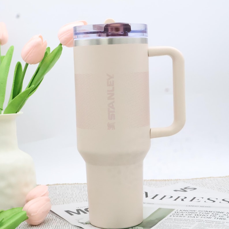Puede incluir: Un vaso t&eacute;rmico Stanley beige con asa y tapa transparente. El vaso tiene una banda texturizada y el logotipo de Stanley. El fondo incluye un jarr&oacute;n con flores rosas y un peri&oacute;dico.
