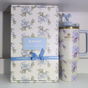 May include: A Stanley Loveshackfancy tumbler and gift box. The tumbler and box are decorated with a floral and bow pattern in shades of blue, pink, and white. The tumbler has a light blue handle and lid. The gift box has a blue ribbon.