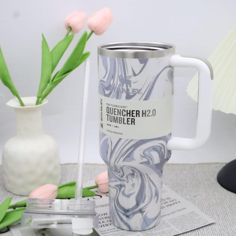 May include: A Stanley Quencher H2.0 tumbler with a white and grey marble design. The tumbler has a white handle and a clear straw. The text "QUENCHER H2.0 TUMBLER" is printed on the side. It holds 1.18L.