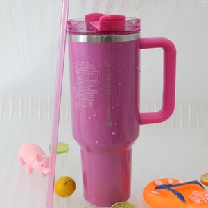 Puede incluir: Un vaso Stanley Starbucks rosa con asa y tapa transparente. El vaso tiene una pajita rosa y el texto "STANLEY x STARBUCKS" impreso en el lateral. El vaso est&aacute; rodeado de un cerdo rosa, un lim&oacute;n y rodajas de lima.