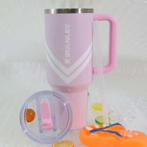 Puede incluir: Un vaso Stanley rosa con un dise&ntilde;o de chevron blanco y rosa y un asa rosa a juego. El vaso tiene una tapa transparente y una pajita transparente. Una chancla naranja y azul est&aacute; en primer plano.