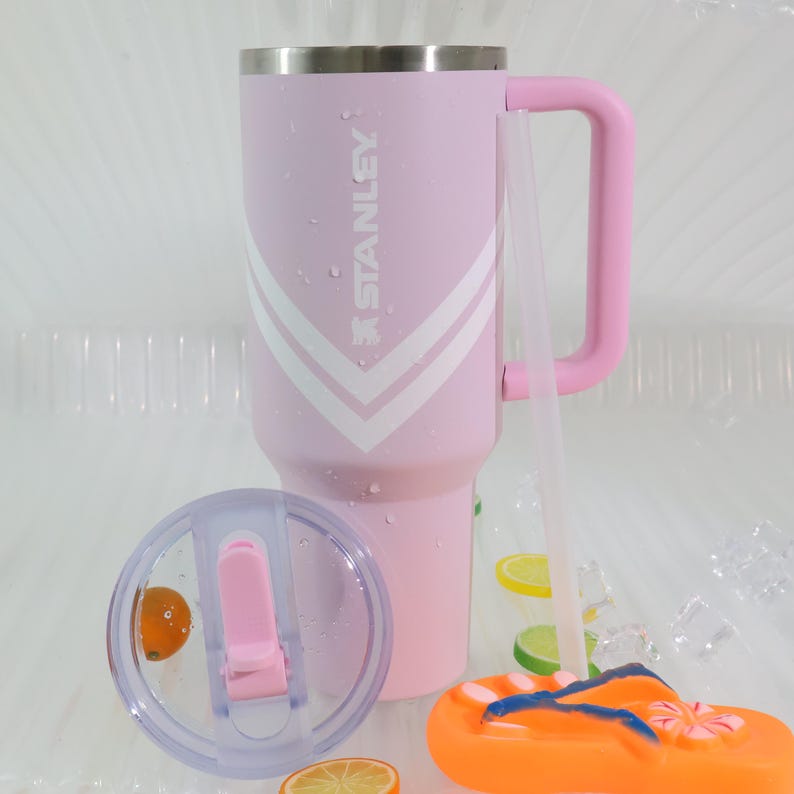 Puede incluir: Un vaso Stanley rosa con un dise&ntilde;o de chevron blanco y rosa y un asa rosa a juego. El vaso tiene una tapa transparente y una pajita transparente. Una chancla naranja y azul est&aacute; en primer plano.