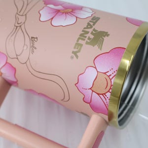 K&ouml;nnte beinhalten: Ein rosa Stanley-Becher mit floralem Design und dem Wort "Barbie". Der Becher hat einen goldenen Rand und Griff. Die Blumen sind rosa und wei&szlig;. Das Stanley-Logo ist goldfarben.