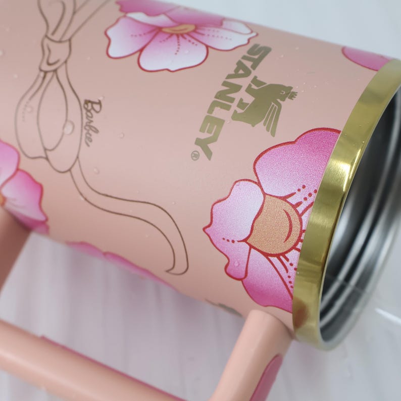 K&ouml;nnte beinhalten: Ein rosa Stanley-Becher mit floralem Design und dem Wort "Barbie". Der Becher hat einen goldenen Rand und Griff. Die Blumen sind rosa und wei&szlig;. Das Stanley-Logo ist goldfarben.