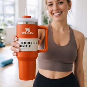 May include: Orange Stanley Quencher H2.0 Tumbler with a white lid and handle. The tumbler has the Stanley logo and product information printed on the front. It is designed to hold 40 oz. of liquid and includes a straw.