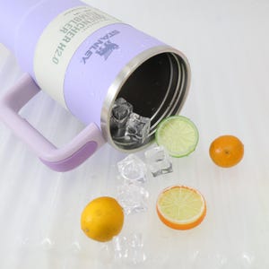 Może przedstawiać: Lawendowy kubek Stanley Quencher H2.0 z uchwytem, ​​obręczą ze stali nierdzewnej i logo Stanley. Kostki lodu i plasterki cytryny, limonki i pomarańczy są rozrzucone.