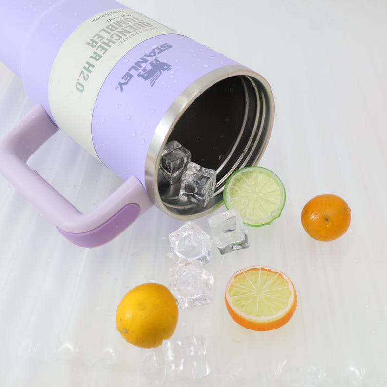 Może przedstawiać: Lawendowy kubek Stanley Quencher H2.0 z uchwytem, ​​obręczą ze stali nierdzewnej i logo Stanley. Kostki lodu i plasterki cytryny, limonki i pomarańczy są rozrzucone.