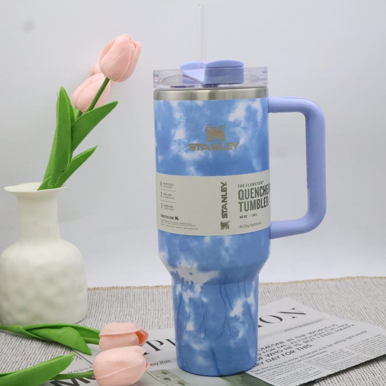 May include: A blue and white tie-dye Stanley Quencher tumbler with a handle and straw. The tumbler has a capacity of 1.18 litres. The tumbler has the Stanley logo and the words "THE FIRST QUENCHER TUMBLER" printed on the side.