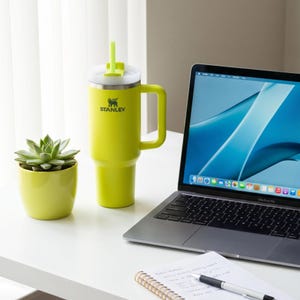 Op de afbeelding: Een limoengroene Stanley-beker met een handvat en een rietje, naast een kleine vetplant in een bijpassende pot. Een laptop en een notitieblok met een pen staan ook op het witte bureau. De beker heeft het Stanley-logo.