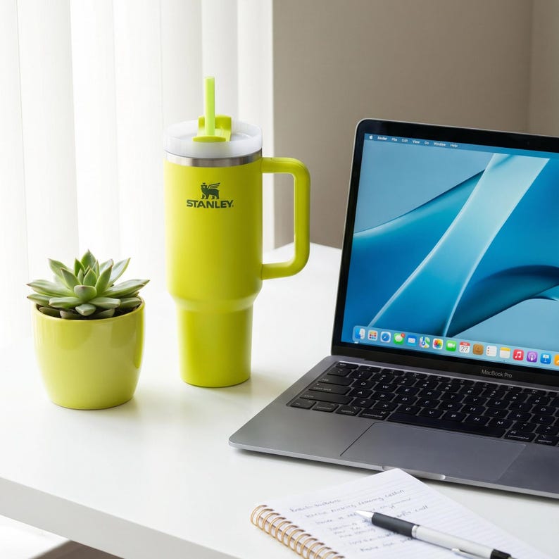 Op de afbeelding: Een limoengroene Stanley-beker met een handvat en een rietje, naast een kleine vetplant in een bijpassende pot. Een laptop en een notitieblok met een pen staan ook op het witte bureau. De beker heeft het Stanley-logo.